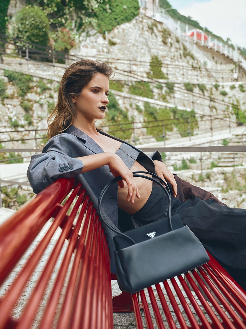A woman in a grey cropped blazer and dark trousers poses reclining on a bright red slatted bench next to a black leather handbag.