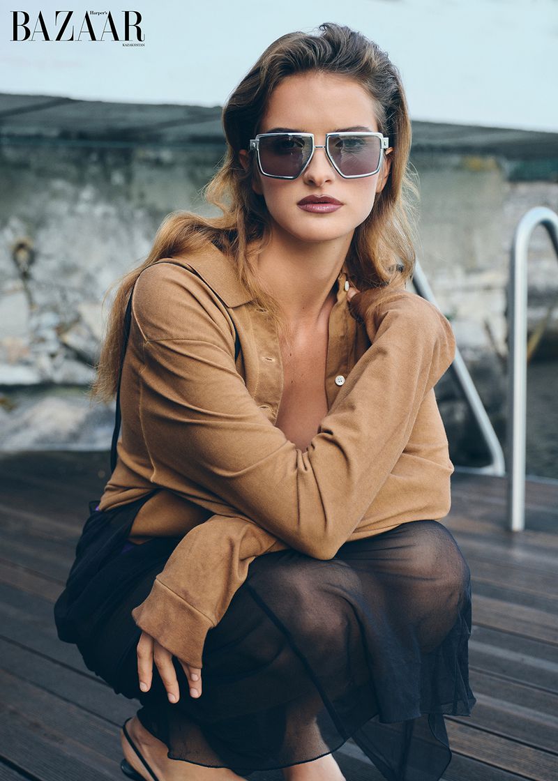Model wearing geometric sunglasses and a tan long-sleeve shirt crouching on a wooden lakeside dock.