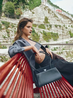 A woman in a grey cropped blazer and dark trousers poses reclining on a bright red slatted bench next to a black leather handbag.