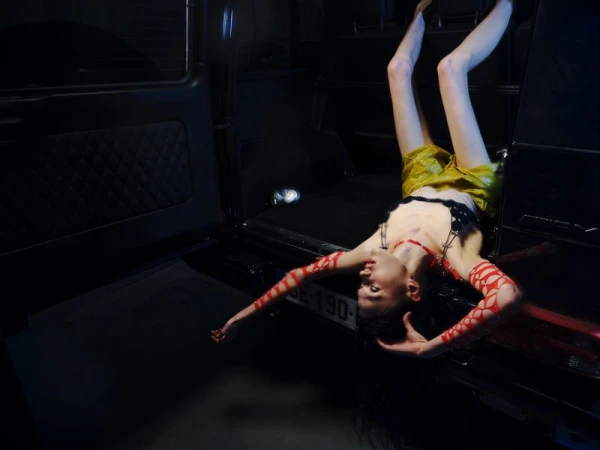 Fashion model posing upside down out of the open trunk of a black SUV wearing yellow shorts and red patterned sleeves.