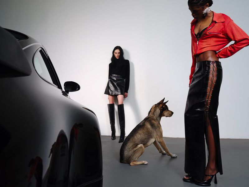 Model in red jacket and black skirt standing next to a dog and a dark car in a studio