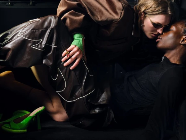 Two models in dark leather garments and sunglasses leaning close together in a dim car interior with a lime green high heel nearby.