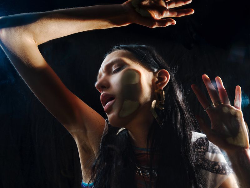 Woman pressing her face and open hands against a clear glass pane in dramatic low-key lighting against a dark background.
