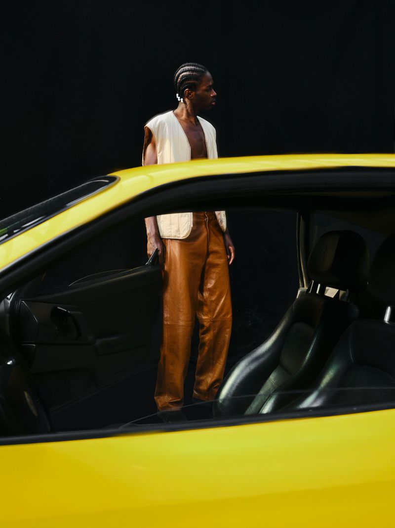 A Black man in a cream vest and brown leather pants stands behind the open window of a bright yellow car against a black background.