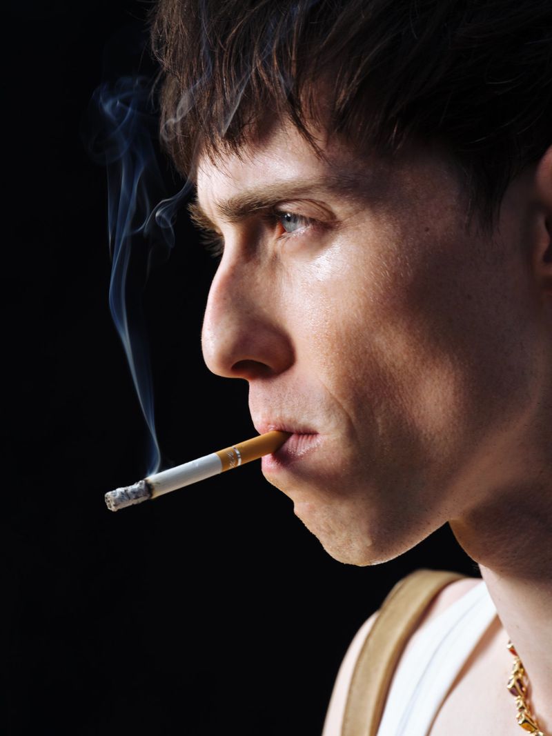 Profile close-up of a man with short brown hair smoking a lit cigarette against a solid black background.