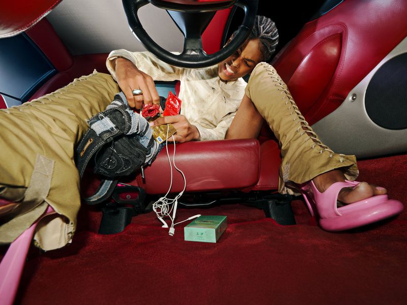 Model in tan lace-up trousers and pink chunky heels sits in a red leather car interior surrounded by scattered items.