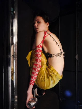 Model crouching in yellow satin pants and red cutout sleeve holding a crushed can against a dark backdrop.