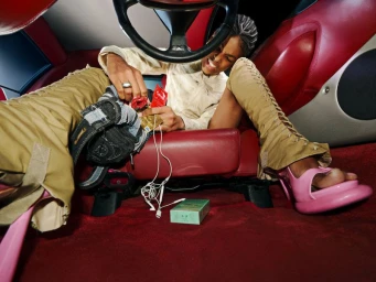 Model in tan lace-up trousers and pink chunky heels sits in a red leather car interior surrounded by scattered items.