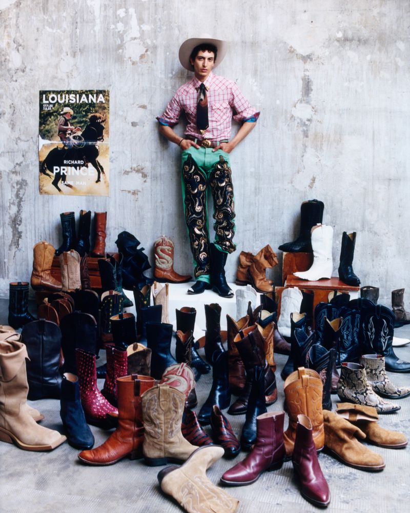 Model Takfarines Bengana in western wear and a cowboy hat standing among dozens of cowboy boots against a concrete wall.