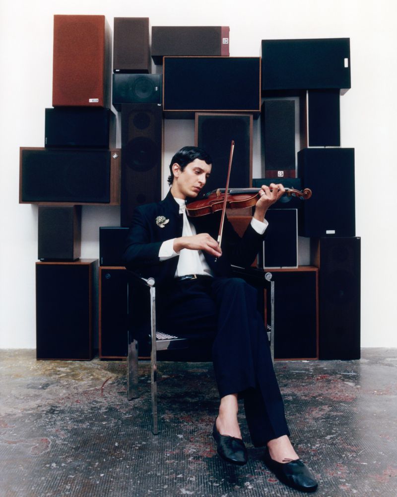 A man seated in a modern chair playing a violin against a wall stacked with various audio speakers.