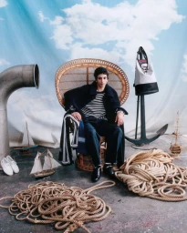 Model Takfarines Bengana seated in a peacock chair surrounded by coiled marine ropes and nautical props before a painted cloud backdrop.