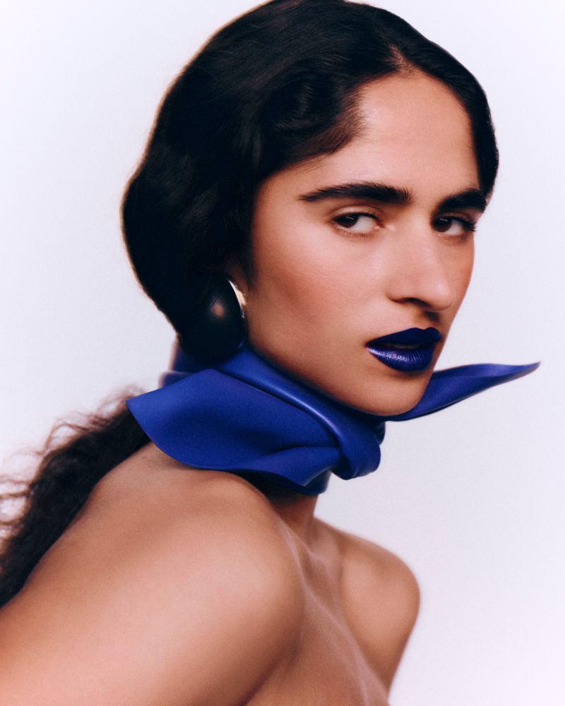 Woman wearing royal blue lipstick and a matching blue leather neck scarf against a clean white studio background.