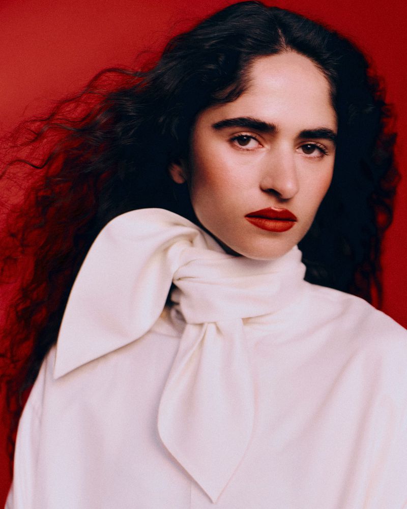 Portrait of a woman with curly dark hair and bold red lips wearing a high-collared white tied blouse against a red background.