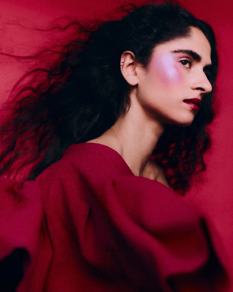 Side profile of a woman with vibrant pink blush, dark lipstick, and wavy hair against a deep red background.