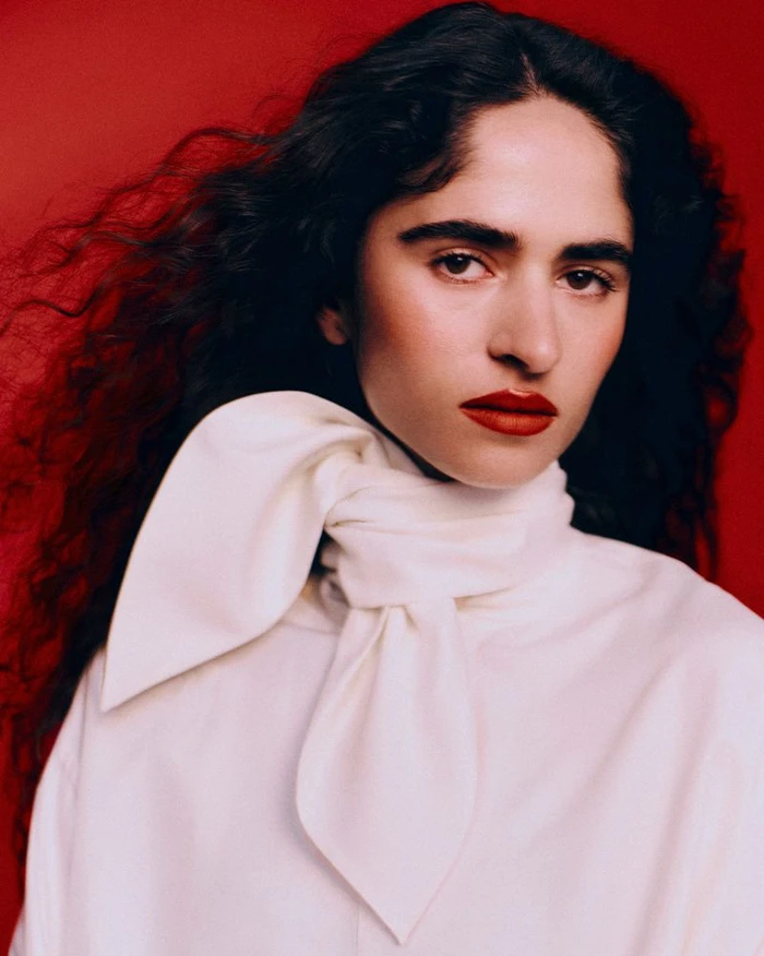 Portrait of a woman with curly dark hair and bold red lips wearing a high-collared white tied blouse against a red background.