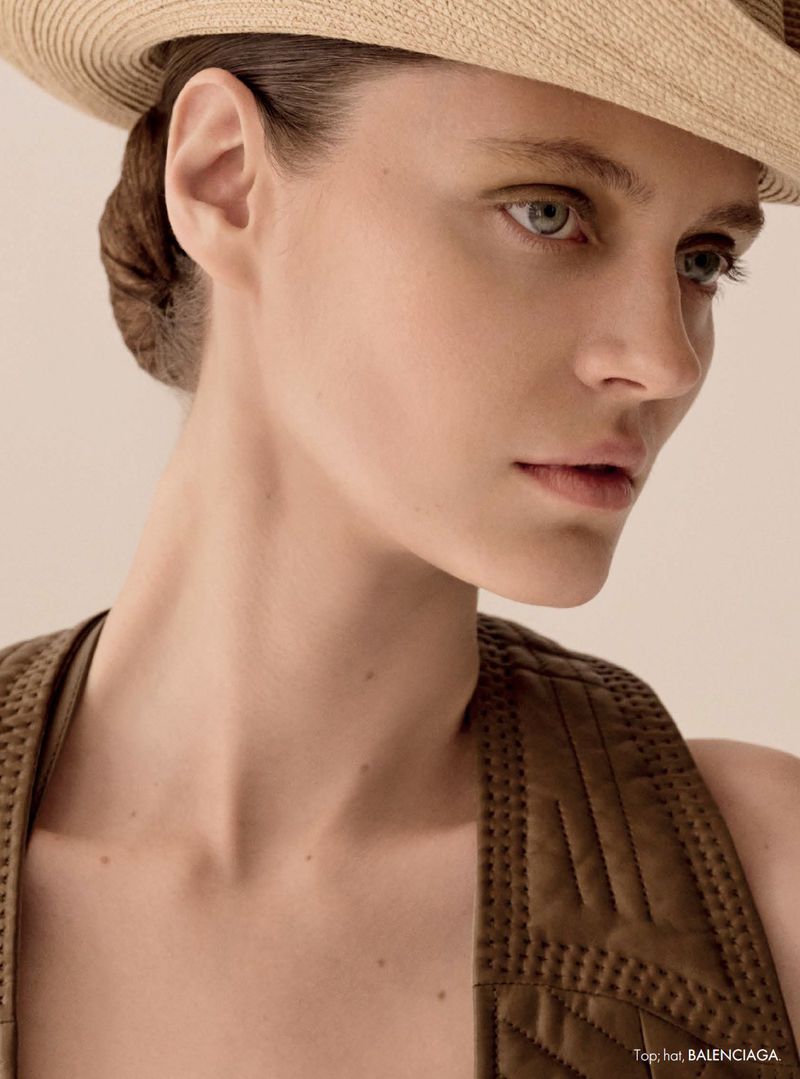 Close-up portrait of model Tali Bulycheva wearing a woven straw hat and a brown quilted top against a warm neutral backdrop.