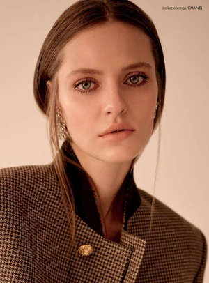 Close-up portrait of model Tali Bulycheva wearing a brown houndstooth jacket and ornate earrings against a cream backdrop.