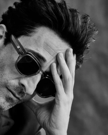 Black and white close-up portrait of actor Adrien Brody wearing dark sunglasses with a hand resting on his face.