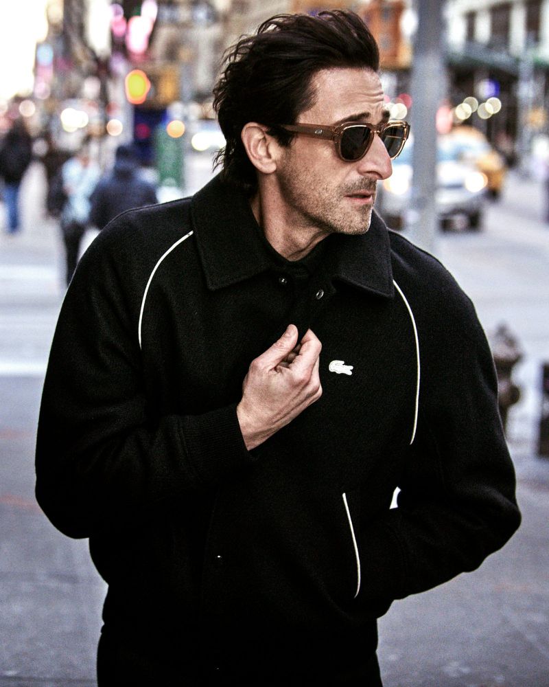 Actor Adrien Brody wearing tortoiseshell sunglasses and a black collared jacket with white piping while walking on a city street.