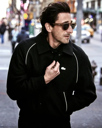 Actor Adrien Brody wearing tortoiseshell sunglasses and a black collared jacket with white piping while walking on a city street.