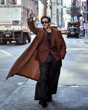 Adrien Brody walks down a city street wearing a long brown coat, blazer, dark wide-leg trousers, and sunglasses with one arm raised.