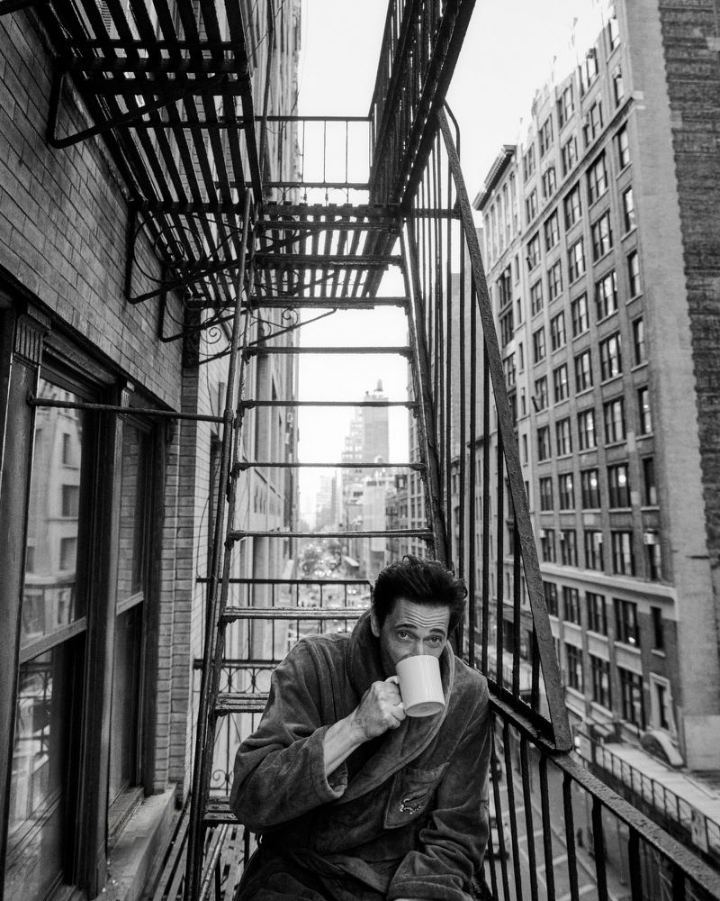 Actor Adrien Brody drinking from a white mug on a metal fire escape overlooking a city street in a black and white editorial photograph.