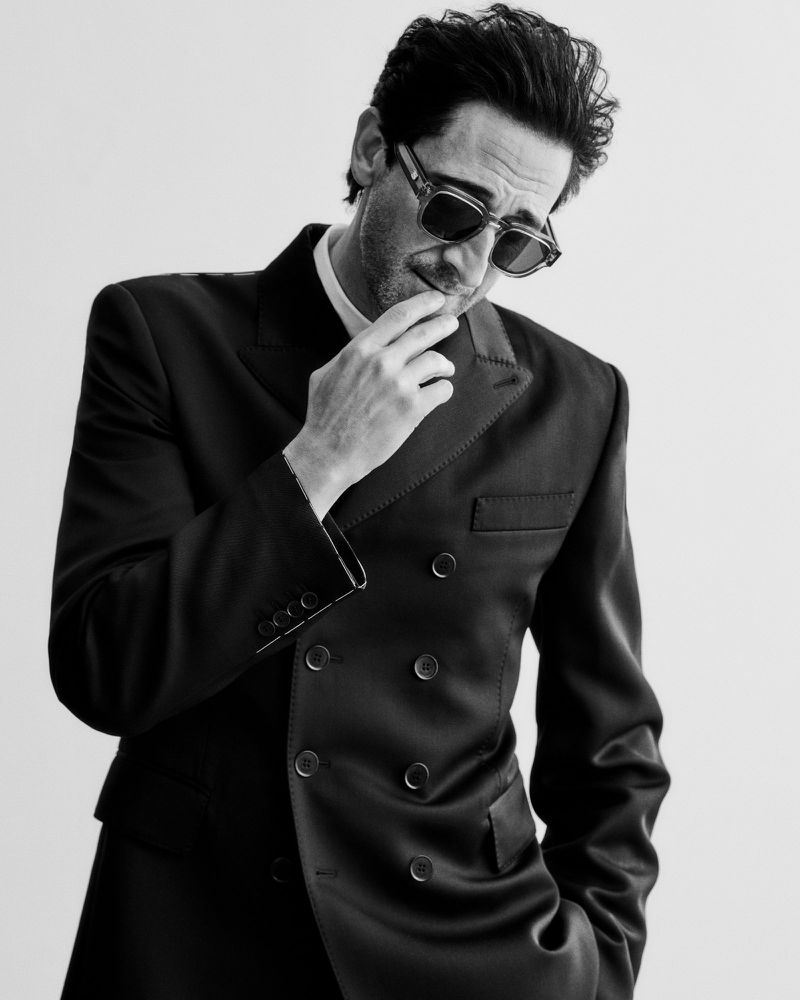 Adrien Brody in sunglasses and a double-breasted black jacket posing with hand to chin against a white studio background.