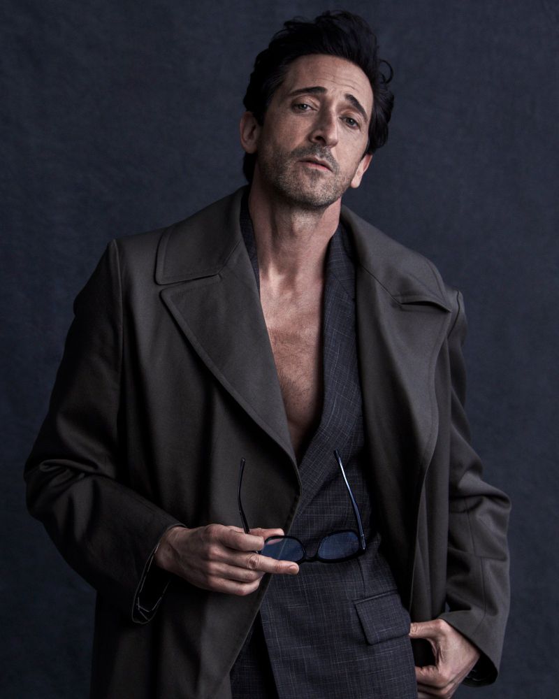 Adrien Brody wearing an open dark trench coat over a grey plaid suit holding sunglasses against a dark studio backdrop.
