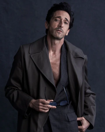 Adrien Brody wearing an open dark trench coat over a grey plaid suit holding sunglasses against a dark studio backdrop.