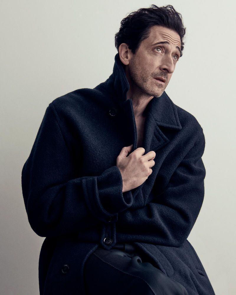 Actor Adrien Brody wearing a dark wool coat while holding the collar against a pale neutral studio backdrop.