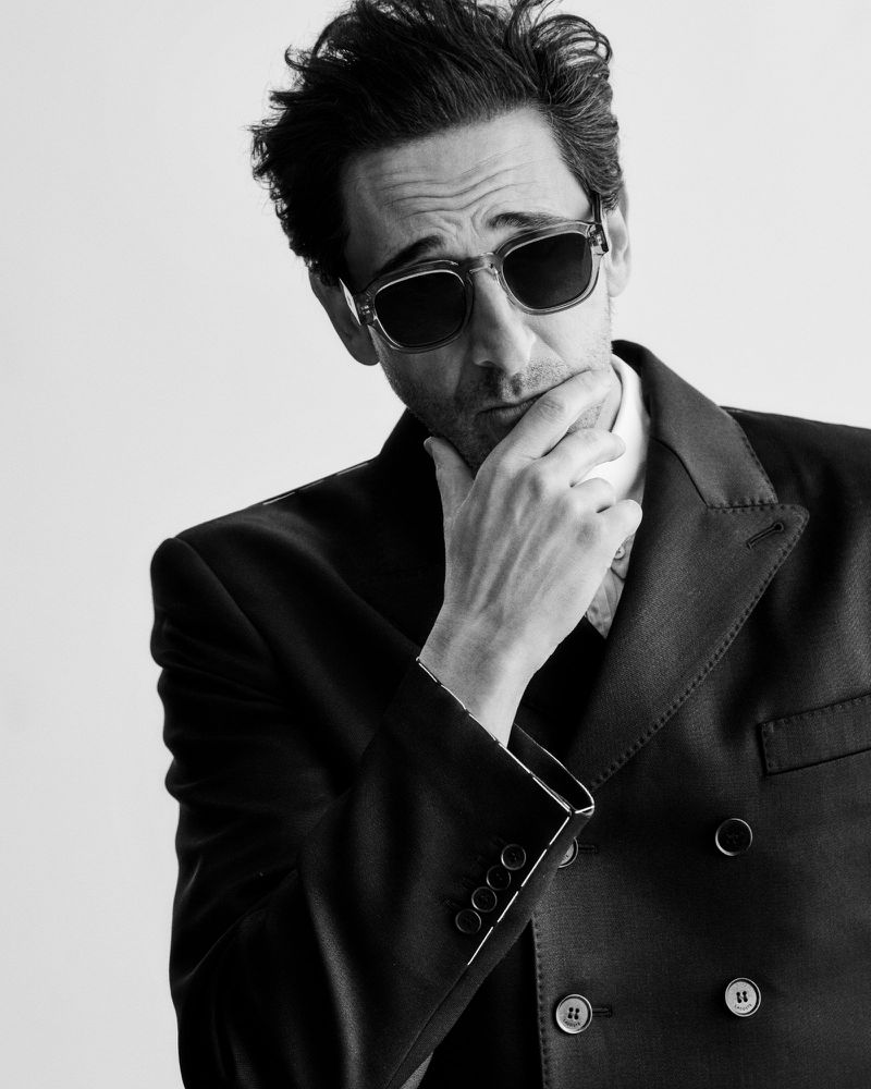 Black and white portrait of actor Adrien Brody wearing sunglasses and a double-breasted jacket against a plain background