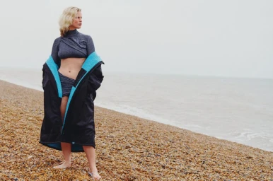 Luca Biggs standing on a pebble beach wearing a grey crop set under an oversized black coat with bright blue lining.