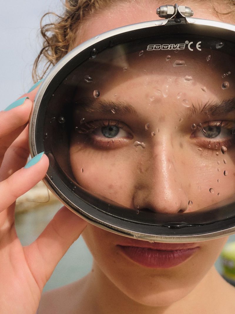 Close-up portrait of a woman looking through wet diving mask, holding it up to her face.