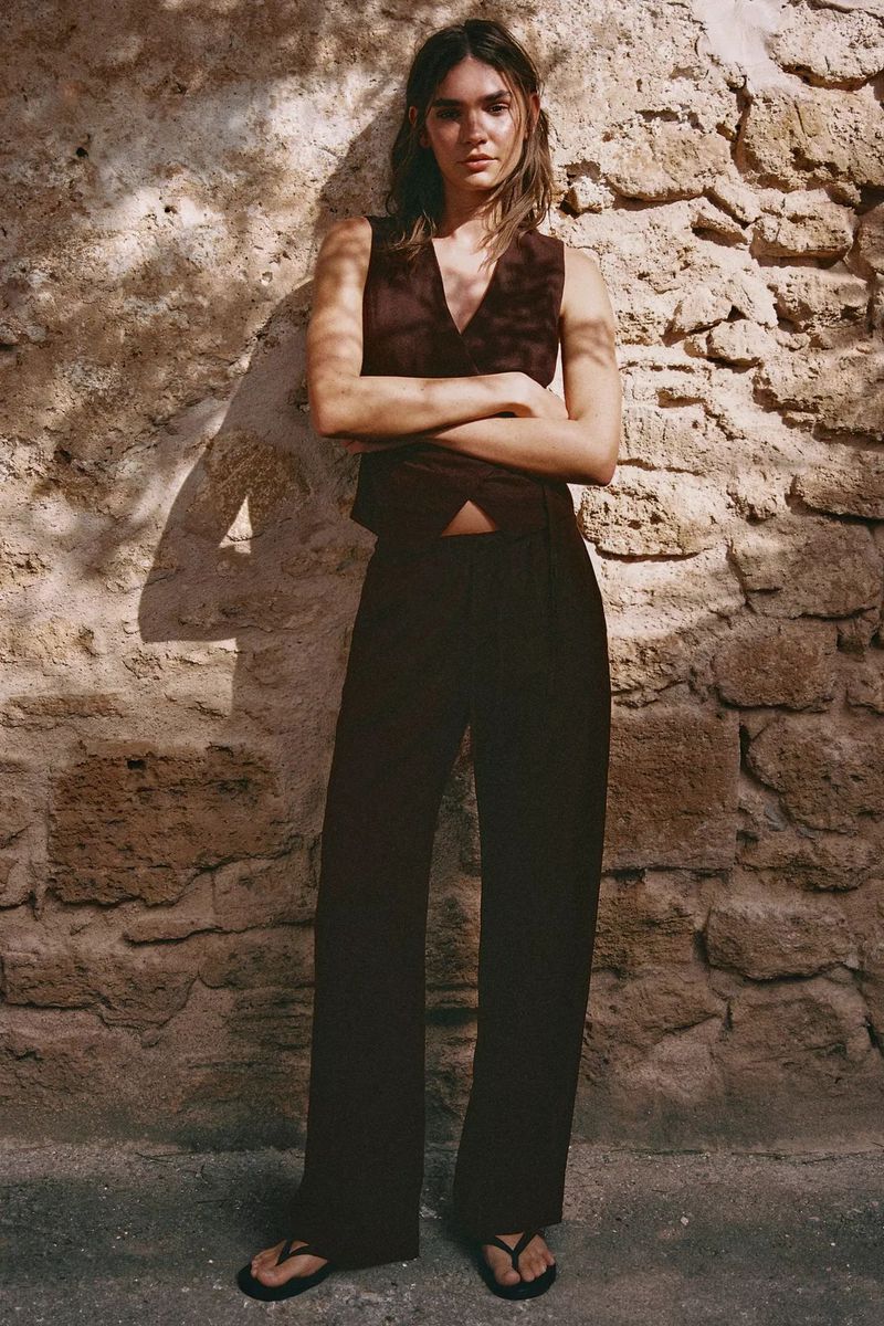 Model standing with crossed arms against a textured stone wall wearing a dark sleeveless vest, wide-leg trousers, and sandals.