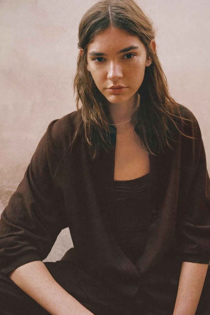 A woman with long brown hair wearing a dark brown button-up shirt sits against a neutral textured backdrop.