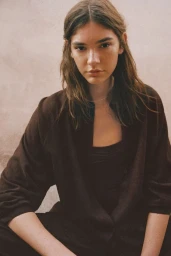 A woman with long brown hair wearing a dark brown button-up shirt sits against a neutral textured backdrop.