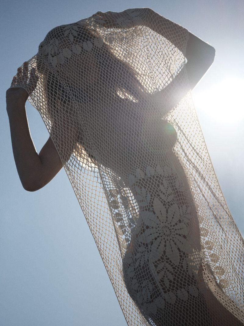 Model holding a sheer cream floral crochet dress over her head backlit by direct sunlight against a pale blue sky.