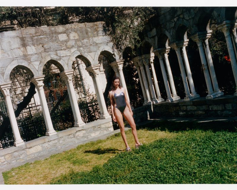 Model Adele Aldighieri posing in a metallic silver one-piece swimsuit on a lawn before stone colonnade arches.