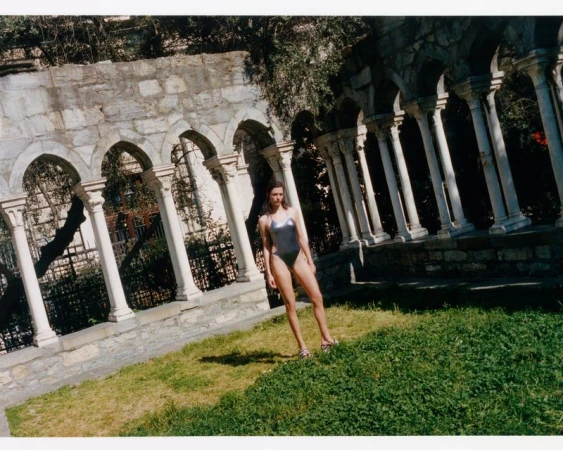 Model Adele Aldighieri posing in a metallic silver one-piece swimsuit on a lawn before stone colonnade arches.