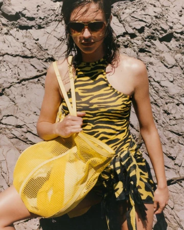 Model wearing a yellow and black tiger print swimsuit, aviator sunglasses, and carrying a yellow mesh bag against a rocky cliff face.