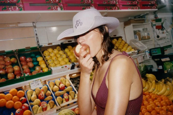 Model Adele Aldighieri wearing a wide brim sun hat and knit top while biting into an apple at a fruit market stall.