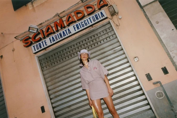 Model Adele Aldighieri in a striped two-piece mini skirt set and hat posing in front of a shuttered Italian storefront.