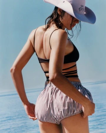 Model Adele Aldighieri wearing a black wrap bikini top, striped shorts, and wide-brim sunhat facing the ocean for The Attico.
