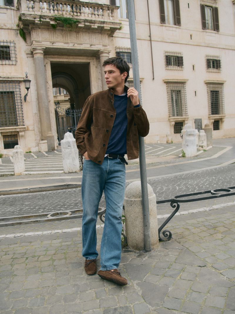 Man wearing a brown suede jacket, blue t-shirt, and blue denim jeans standing on a cobblestone European street.
