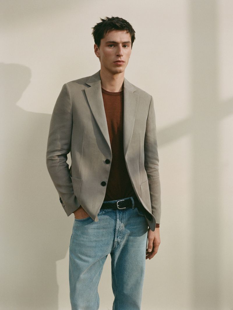 Model Clément Laguardia wearing a grey tailored blazer, brown knit shirt, and light wash denim jeans against a neutral studio wall.