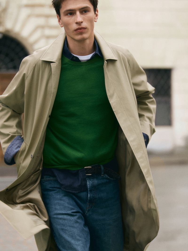 Clément Laguardia wearing a beige trench coat, green knit sweater, and blue denim jeans on an urban street.