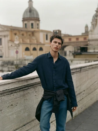 Model Clément Laguardia in a navy button-down shirt and blue jeans leaning against a stone balustrade with historic city domes behind.