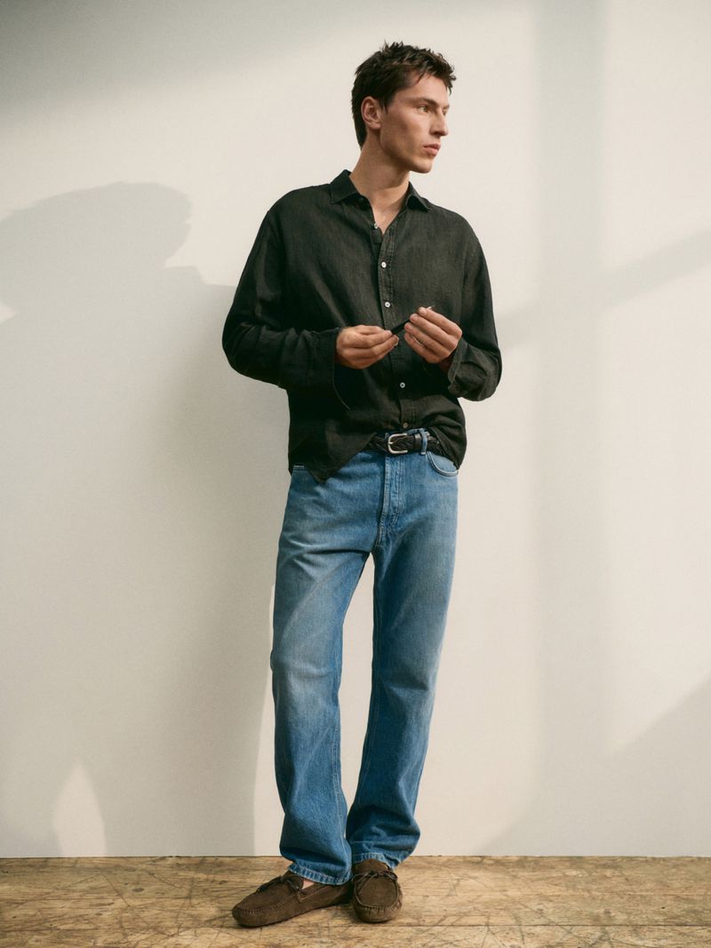 Model Clément Laguardia wears a dark button-down shirt and blue denim jeans against a neutral studio backdrop with window shadows.
