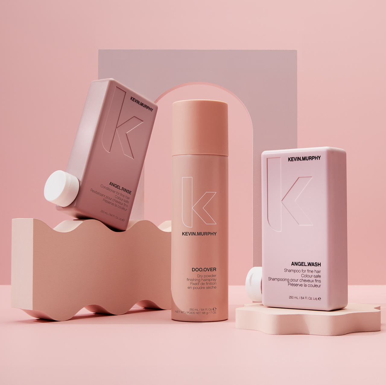 Still life shot of three Kevin Murphy hair product bottles in shades of pink, styled with geometric shapes on a pink background.