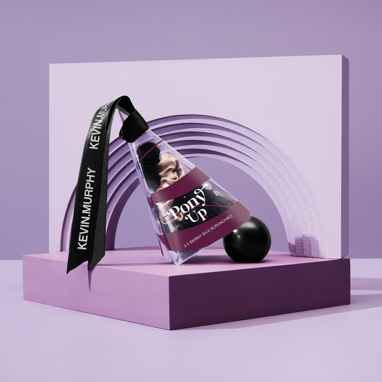 Product shot of Kevin Murphy Pony Up hair scrunchies in clear tube on a purple platform against a lilac backdrop.